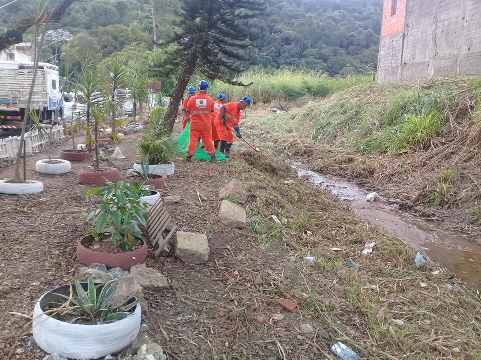 À esquerda, pneus utilizados como vaso de árvores. 4 funcionários ao centro, um com enxada fazendo limpeza do córrego, todos de uniforme laranja e capacete azul.